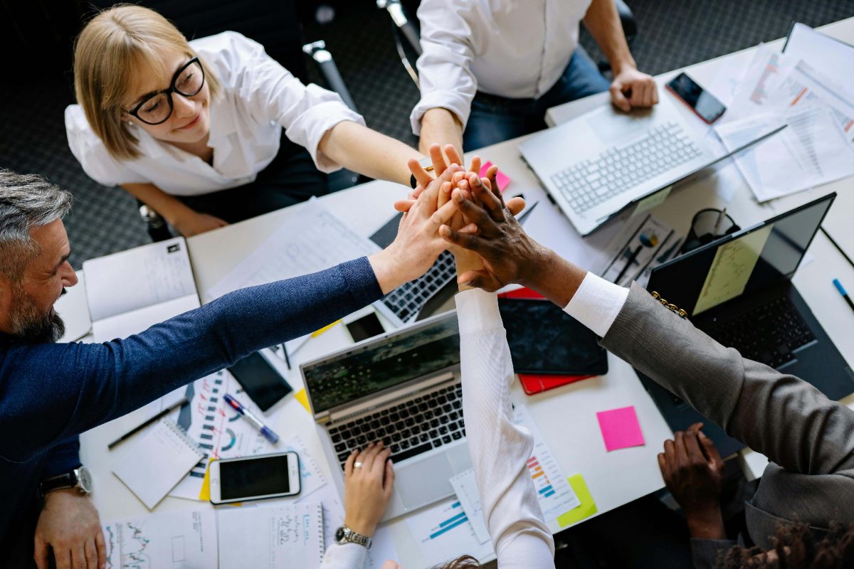Team stacking hands together over a desk with laptops
